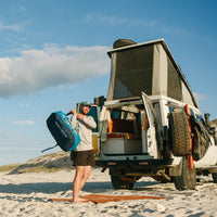 A man puts a blue duffle bag on his shoulder while standing next to a camper van on the beach