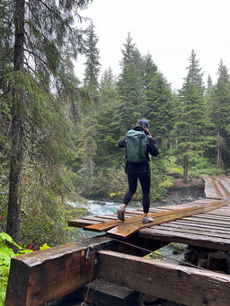 Person with a green Eagle Creek backpack crossing a wooden bridge over a forest stream
