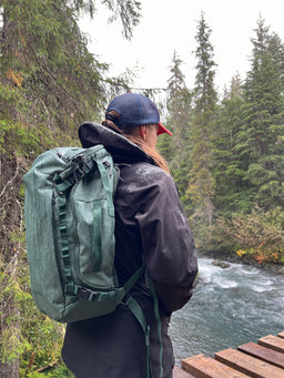 Person with a green Eagle Creek backpack standing by a river in a forest
