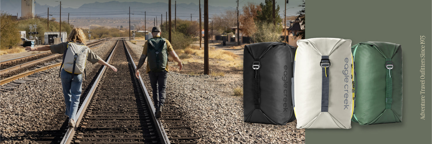 Two people walk down railroad tracks wearing their Eagle Creek Tour Travel Backpacks