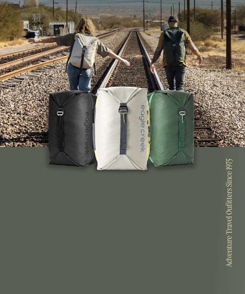 Two people walk down railroad tracks wearing their Eagle Creek Tour Travel Backpacks