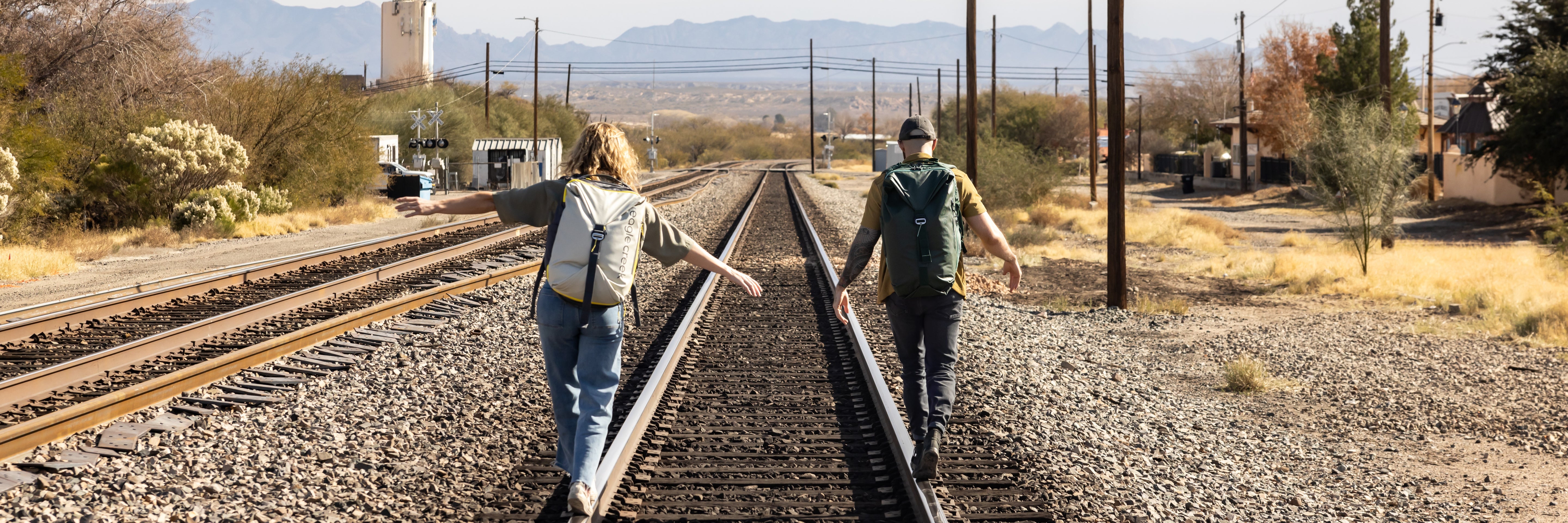 Two people walk along railroad tracks wearing Eagle Creek travel backpacks