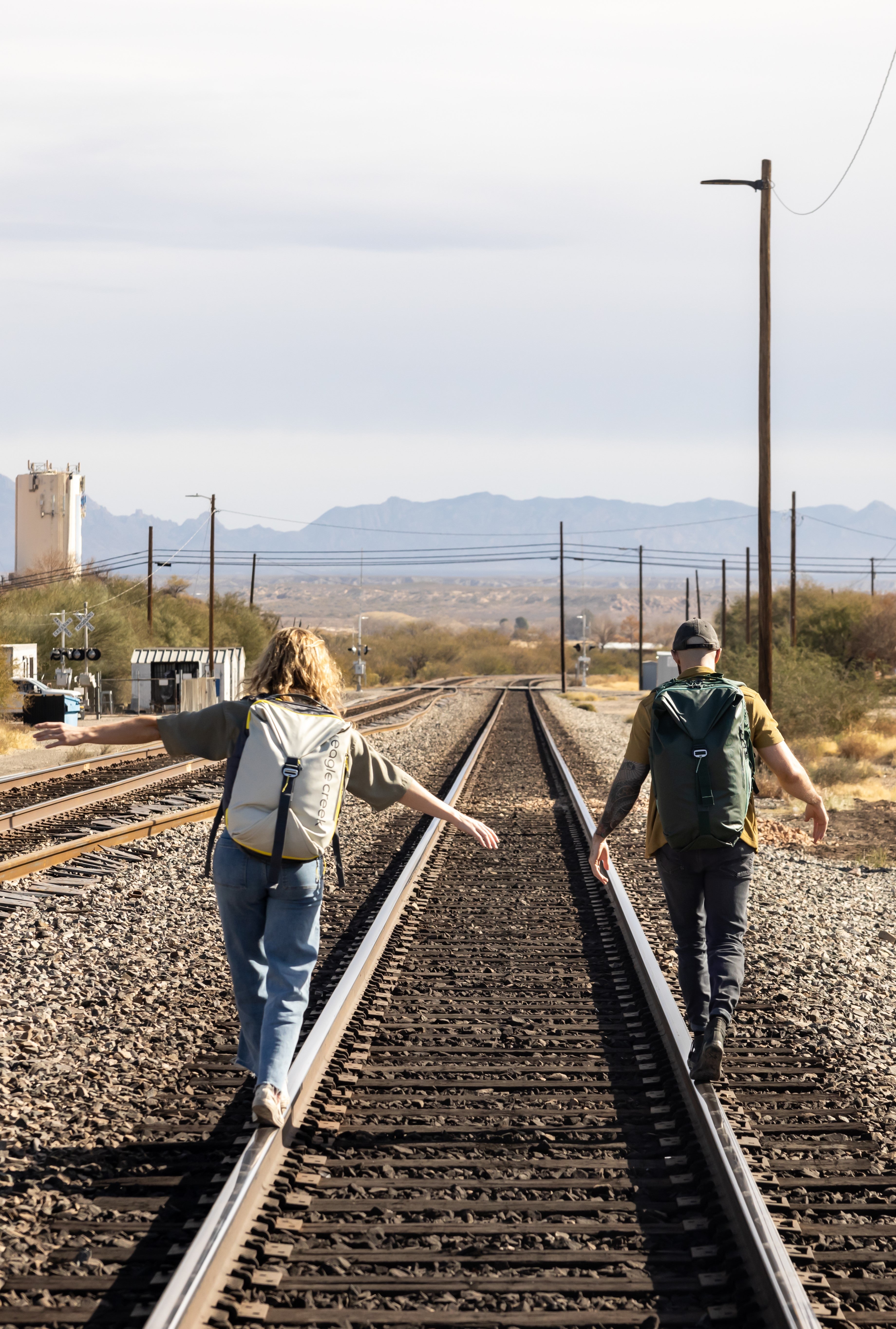 Two people walk along railroad tracks wearing Eagle Creek travel backpacks